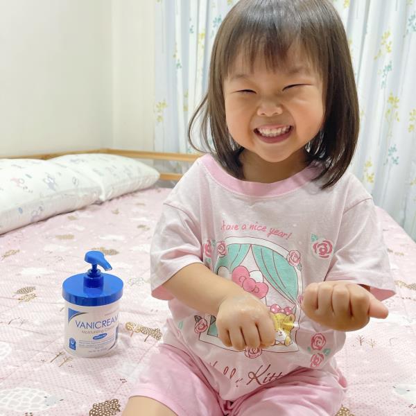 三寶媽Claire育兒札記拍攝小朋友笑容滿面的坐在床上，周圍有一瓶薇霓肌本全日高效修護保濕乳霜家庭號