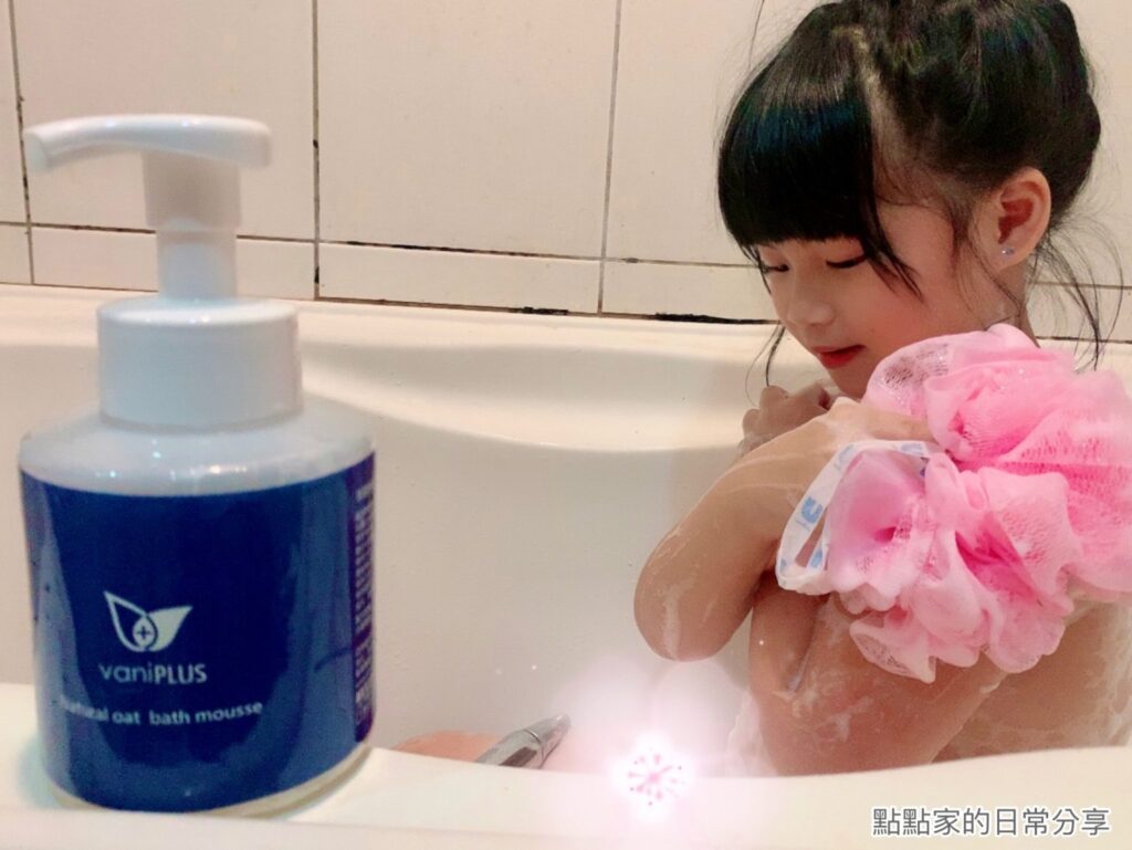 點點家的日常分享拍攝點點小妞坐在浴缸裡，手上拿著浴球搓正在洗身體，周圍有一瓶薇霓進階天然燕麥潔膚慕斯