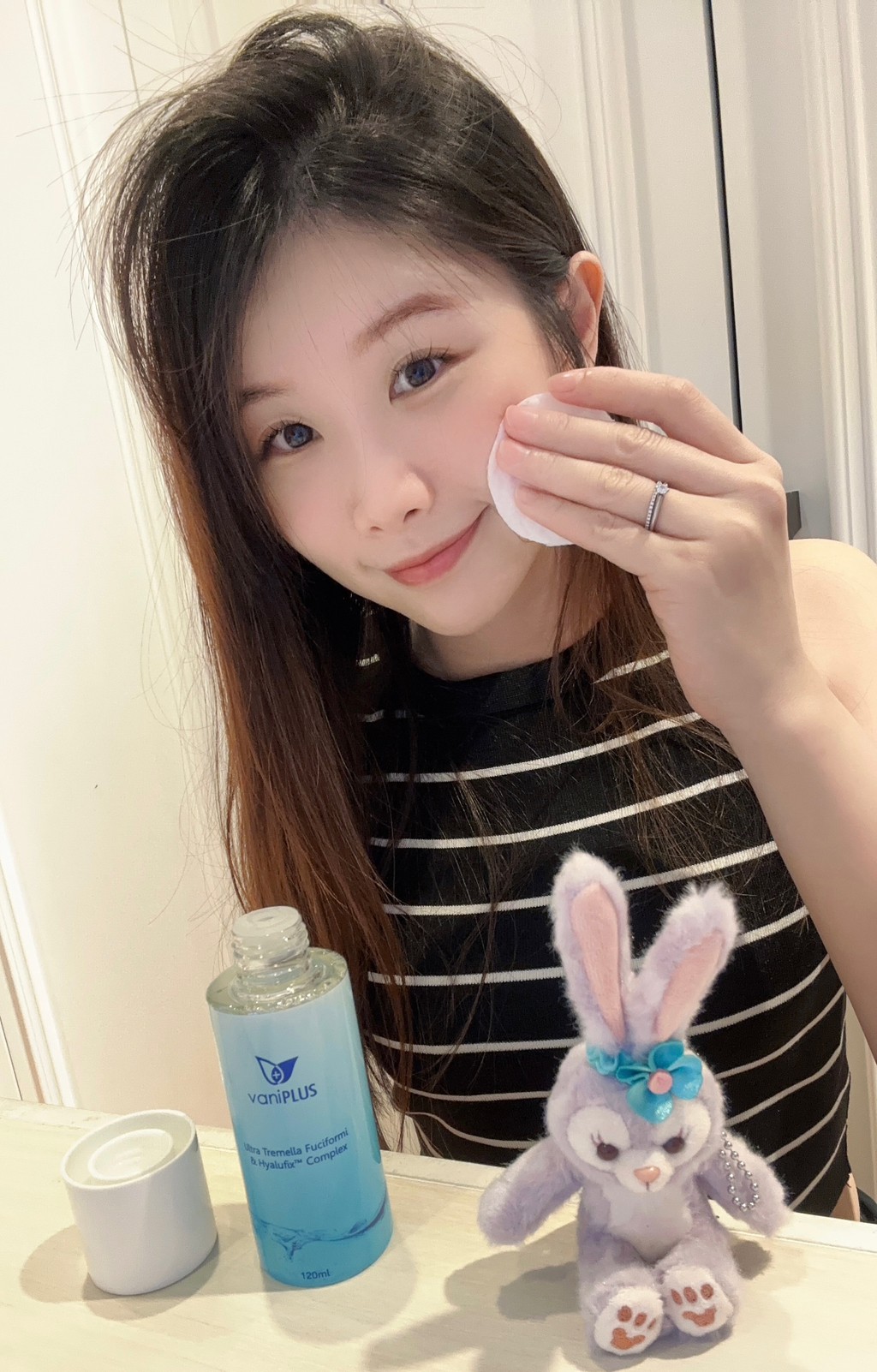 薇琪の萬得窩vicky將手中的化妝棉用薇霓進階極潤雪耳精露沾濕之後擦在臉頰肌膚上，周圍有一隻史黛拉兔娃娃