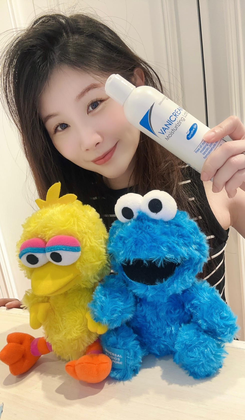 薇琪の萬得窩拍攝vicky坐在米色的桌面前右手拿著薇霓肌本無油安膚清爽調理乳液拍合照，周圍有一隻Cookie Monster與Big Bird娃娃