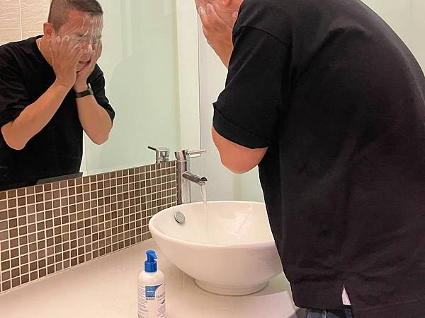 Wu’shouse 三個寶包日常拍攝阿V的先生在洗手台前面洗臉時的情境照，周圍有一瓶薇霓肌本胺基酸調理潔膚露