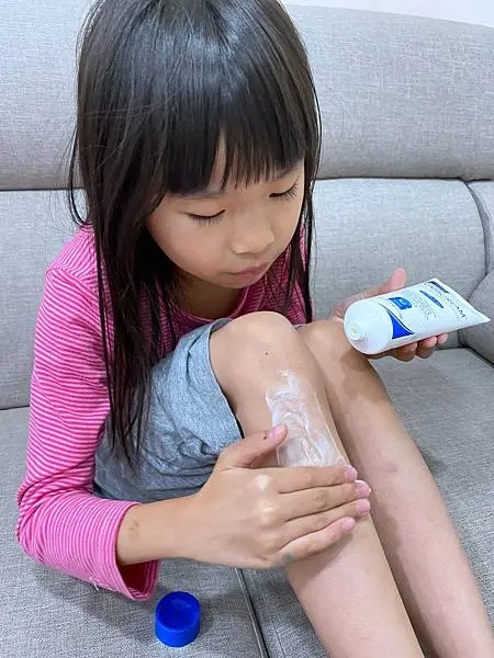 小女孩正在仔細地將VANICREAM薇霓肌本全日高效修護保濕乳霜塗抹在自己的小腿皮膚上，顯示出她正在進行保濕護理。旁邊的沙發上放著藍色的乳霜瓶蓋。