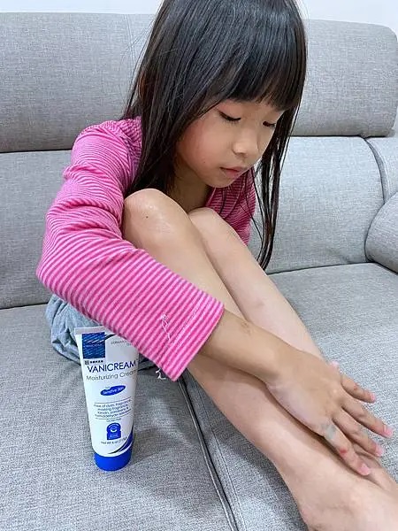小女孩正在仔細地將VANICREAM薇霓肌本全日高效修護保濕乳霜塗抹在自己的小腿皮膚上，顯示出她正在進行保濕護理，背景為淺灰色的沙發，沙發上有細密的紋理。