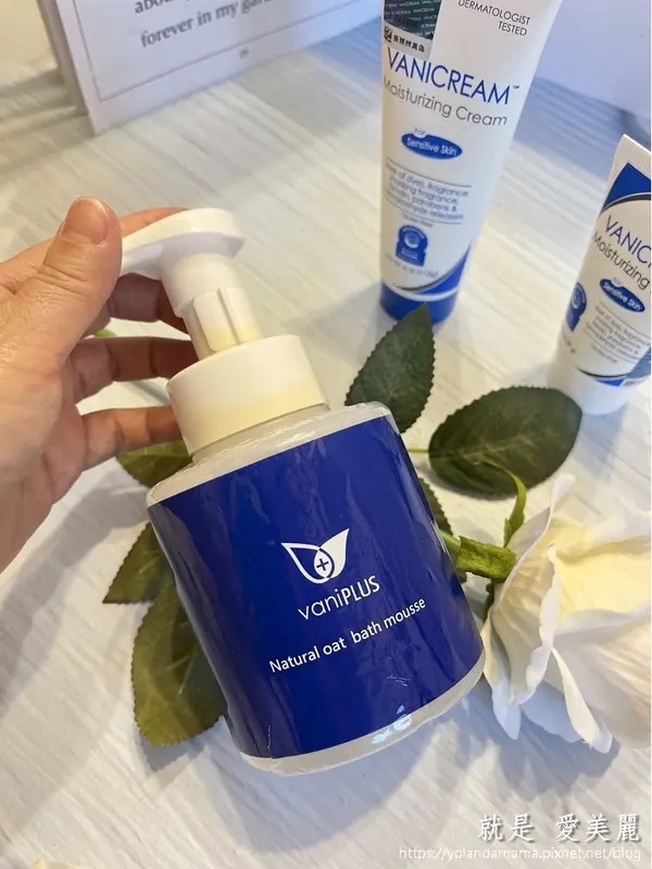 這是一張展示vaniPLUS薇霓進階天然燕麥潔膚慕斯和VANICREAM保濕系列產品的照片，手持的沐浴慕斯是主要焦點，背景有書籍和花葉裝飾。