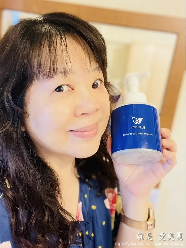這是一張中年女性展示vaniPLUS薇霓進階天然燕麥潔膚慕斯的自拍照，旨在推廣這款產品。笑容和產品的清晰展示都傳達了對產品的喜愛和推薦。