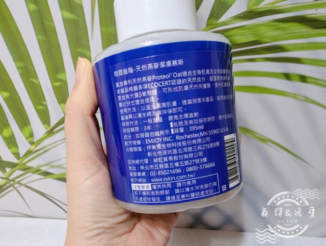 手持薇霓進階天然燕麥潔膚慕斯產品瓶身背面，標示成分來源、容量與使用方式，背景為熱帶綠葉設計