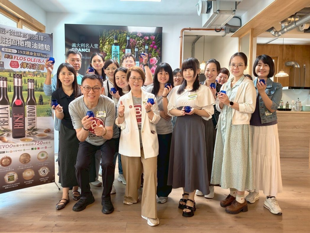 義大利莊園橄欖油體驗活動大合照。多位參與者在室內手持藍色品油杯微笑合影,前方女性雙手比讚。背景展示 FRANCI 名人堂活動海報與 BELLA 等頂級橄欖油品項介紹。