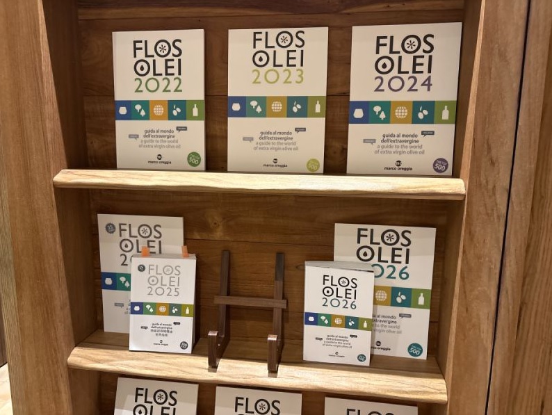 木質書架陳列Flos Olei歷年世界特級初榨橄欖油評鑑專書。上層整齊擺放2022、2023與2024年指南,中層陳列2025與2026年版本與木質書架,展現權威橄欖油品鑑榮譽。