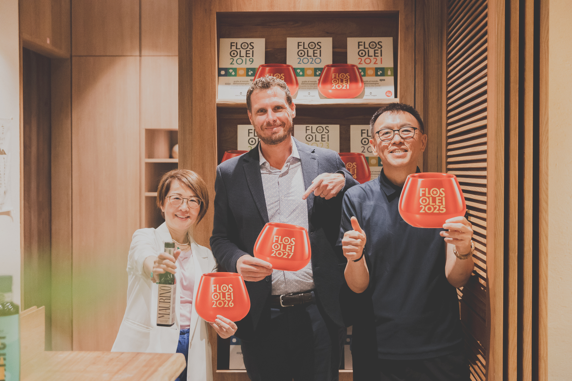 義大利品油師與SUSU姐跟品味大叔手持Flos Olei紅色品油杯與Franci特級初榨橄欖油合影。背景為Flos Olei得獎指南展示櫃，展現頂級莊園橄欖油專業品鑑與登台風采。
