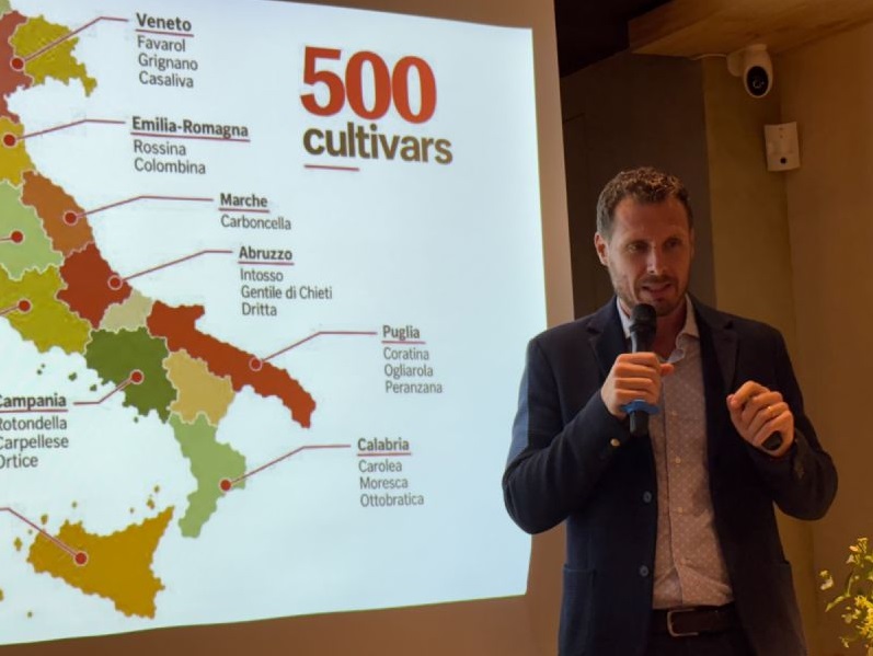 官方認證品油師Federico Ferroni手持麥克風演說。背景投影展示義大利各產區地圖與500 cultivars橄欖品種簡報,深入解析義大利原生橄欖品種多樣性與產區風土特性。