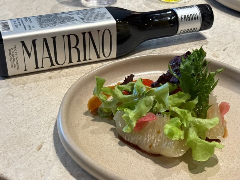 義大利Franci莊園Maurino單一品種特級初榨橄欖油瓶身,平放於精緻鮮蔬沙拉盤旁,盤內盛裝新鮮綠生菜與多汁果肉,呈現頂級橄欖油佐餐搭配與美食饗宴。