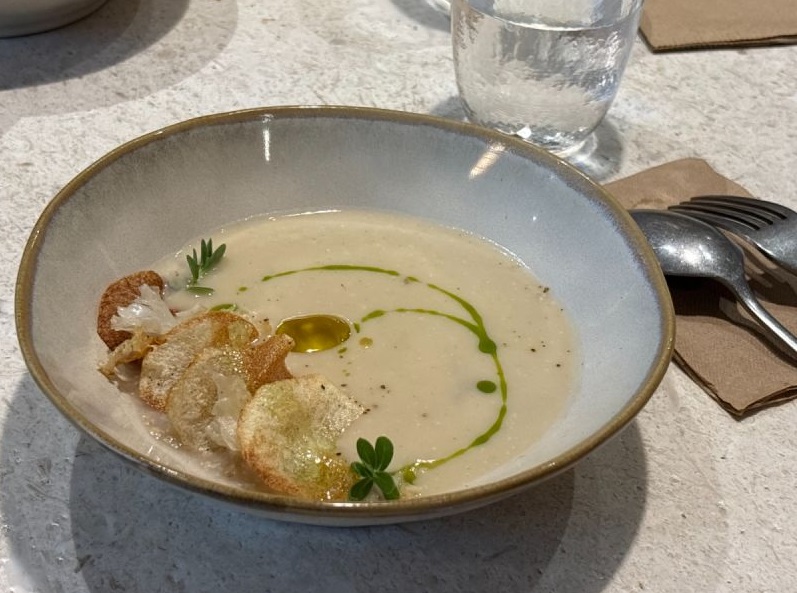 頂級初榨橄欖油佐香濃濃湯料理。陶碗盛裝溫熱濃湯,淋上金黃橄欖油滴與青綠油醋弧線拉花,邊緣點綴香脆洋芋脆片與綠色香草,展現莊園橄欖油精緻佐餐饗宴。